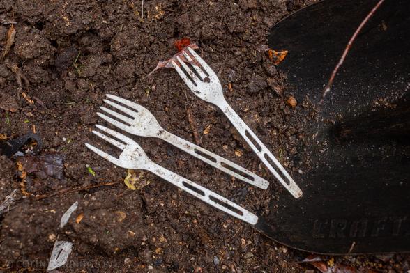 Three dirty white plastic forks, one with a tine missing, on wet soil near the blade of a shovel.