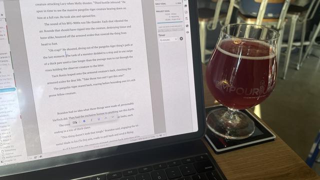 The image shows a laptop on a wooden table with a document open on the screen, displaying a scene from a story involving a creature attacking a character named Lucy. The text is in English and includes dialogue and action descriptions. To the right of the laptop, there is a glass filled with a reddish liquid, likely a craft beer, placed on a coaster with the text "EMPOURRIUM” visible. The background features metal chairs and a blurred view of a café setting. The laptop screen also shows a word processing application with formatting options visible on the right side.