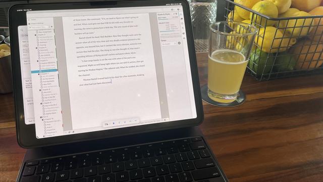 The image shows a tablet on a wooden table, displaying a document with text. The tablet is connected to a black keyboard case, suggesting it is being used as a laptop. The document appears to be a written piece, possibly a story or an article, with visible text and formatting options on the screen. The left side of the screen shows a sidebar with a file directory, indicating the document is part of a larger project or collection. The right side of the screen shows the document's content, with a visible cursor and formatting tools.

In the background, there is a glass of a light-colored beverage, possibly beer, on a coaster. To the right of the tablet, there is a wire basket filled with lemons and limes, adding a touch of color and context to the setting. The overall scene suggests a casual, comfortable work environment, possibly at home or in a relaxed workspace.