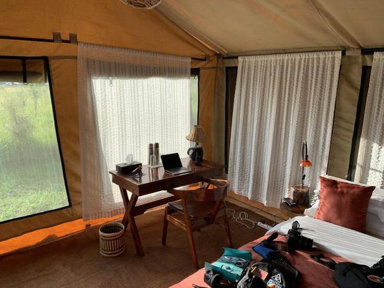 The image depicts the interior of a tent with a cozy and well-organized setup. The tent features a large window with white lace curtains, allowing natural light to fill the space. The floor is covered with a brown carpet, and the walls are made of a beige canvas material.

In the foreground, there is a wooden desk with a laptop, a lamp, and a tissue box, suggesting a workspace. A wooden chair is positioned in front of the desk. To the right, a bed with white linens and a rust-colored pillow is visible, with various items, including a camera and a backpack, placed on it.

A small round table with a lamp is situated next to the bed, and a woven basket is on the floor near the desk. The overall ambiance is warm and inviting, with a mix of natural and modern elements.