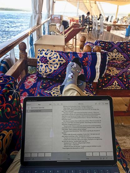 The image shows a person sitting on a colorful, patterned couch on a boat, with their feet up on the seat. They are wearing patterned socks with cartoon characters and beige pants. A laptop is open on their lap, displaying a document titled "Agent Bennise, Space Spy" with the text "Nothing Scary Here" and 306 words. The background features a view of the water and a dining area with tables and chairs. The boat has a wooden railing and a canopy providing shade. The scene is bright, suggesting it is daytime.

Provided by @altbot, generated privately and locally using Ovis2-8B

🌱 Energy used: 2.507 Wh