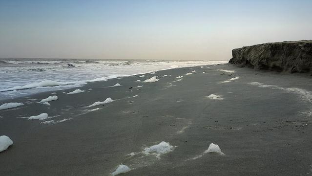 A wide shot of a calm beach with gentle waves meeting the shore. Patches of white seafoam are scattered across the dark sand leading towards a high, steep bank of sand on the right side, showing evidence of recent erosion and collapse. The sky is overcast and a light grey.