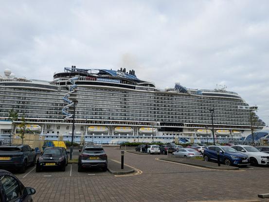 An enormous modern cruise liner, about a dozen decks tall, is moored next to a mundane looking car park.

There is a complex set of tubes running vertically between many of the ship's decks.