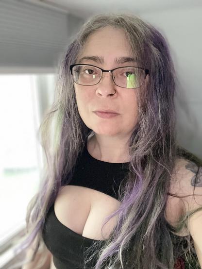 A person with long, slightly wavy hair featuring purple highlights, wearing glasses and a black sleeveless top, poses in front of a window. Soft natural light illuminates the scene.