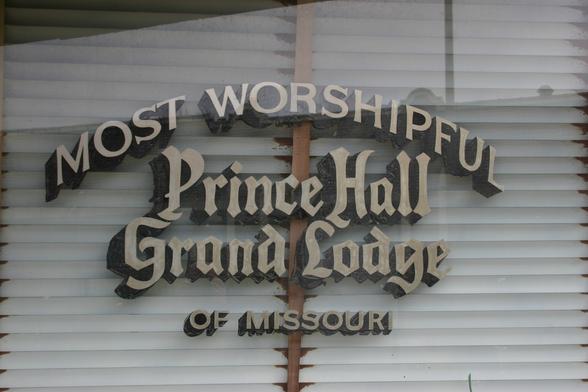 A color photo bearing a sign for a Masonic lodge. The name of the lodge, Prince Hall Grand Lodge, is in a blackletter style, with "Most Worshipful" in an all-caps serif font arced above. Below and smaller is "of Missouri". There are white Venetian blinds, closed, behind the window.