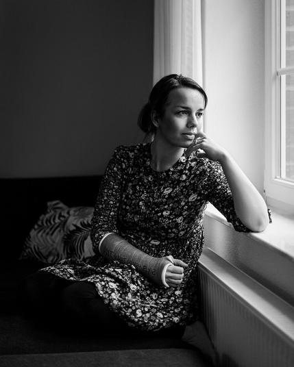 Eine Frau, die aus dem Fenster schaut. Sie sitzt dabei auf einem Teil einer Couch, hat ihren linken Arm auf die Fensterbank gestützt und den rechten Arm, an dem sie einen Gipsverband trägt, auf den Schoß gelegt.