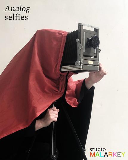 A hooded figure underneath a dark cloth and crouching behind a large format camera. The darkcloth is black on the underside, but a striking red colour on the top.  

The text on the top left reads "Analog selfies" and there is a logo for studio MALARKEY in the bottom right.
