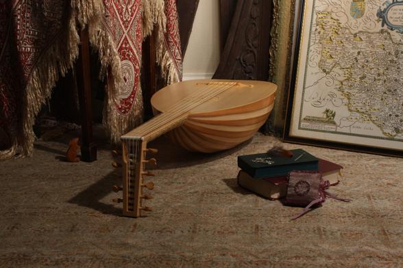 A photograph of a renaissance lute made to look like a 16th century still life painting.

A 6-course early renaissance lute with a striped yew bowl, light fingerboard and heart shaped tuning pegs is lying on the floor. It seems discarded in a corner between a chair draped with with a blanket, patterned in red jacquard, and a stack of old picture frames, the frame in the foreground contains a hand drawn map of the West Riding from 1612. A few books and other personal items are lying on the floor next to the lute. A wooden mouse looks up the chair leg with a curious gaze.