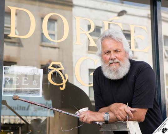 Portrait of a man posing in front of a sign he is painting on a window. He is holidng his sign painting materials in his hands.