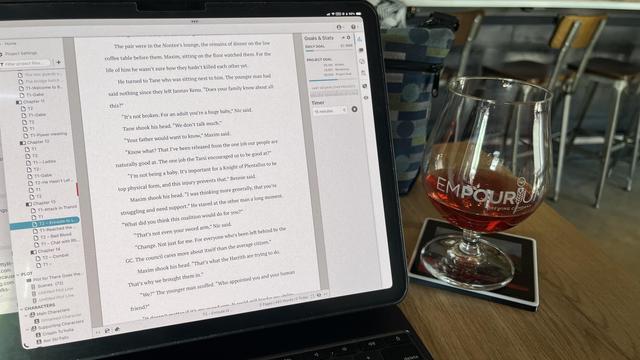 The image shows a close-up of a laptop screen displaying a document, likely a novel or screenplay, with a visible table of contents on the left side. The text on the screen is in English and includes dialogue between characters named Tane and Maxim. The document is open in a word processing application, with a sidebar showing project settings and statistics. The laptop is placed on a wooden table, and next to it is a glass with a reddish-brown liquid, possibly a beverage, on a coaster with the word "EMPOURIUM" printed on it. In the background, there are blurred chairs and a bag, suggesting a casual setting, possibly a café or a co-working space.

Provided by @altbot, generated privately and locally using Ovis2-8B

🌱 Energy used: 0.160 Wh