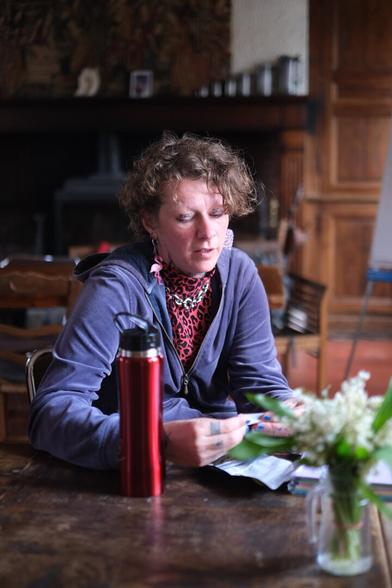 Baxter assis à dans un salon à une table en bois. Ses cheveux sont blonds bouclés, il porte une veste bleue, un haut léopard rose et des boucles d'oreilles flamands roses. il roule une cigarette. Sur la table il y a une gourde rouge et un bouquet de muguets.