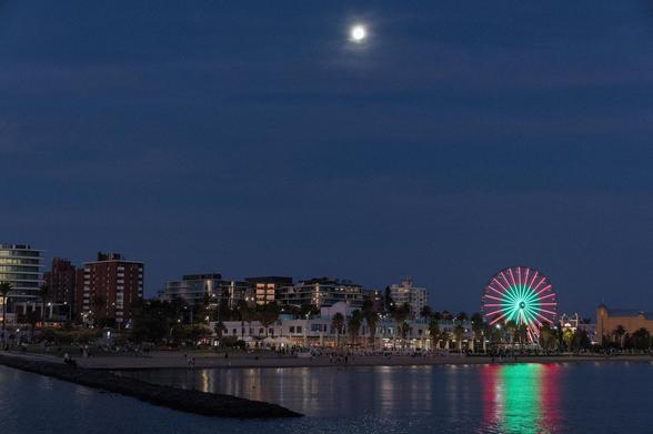 A landscape photo taken at the end of the day at sunset on a beach. The view is directed towards the beach. A few high-rise buildings can be seen on the promenade, their windows reflecting the light of the sunset. There is a large Ferris wheel on the beach, illuminated in two rings - the outer one in red, the inner one in green. The light is broadly reflected on the surface of the calm water. Above the scene is the almost full moon and a few light clouds.

AI disclaimer: Using my work, its meta data, written or derived description to create media with or train AI based systems is prohibited.