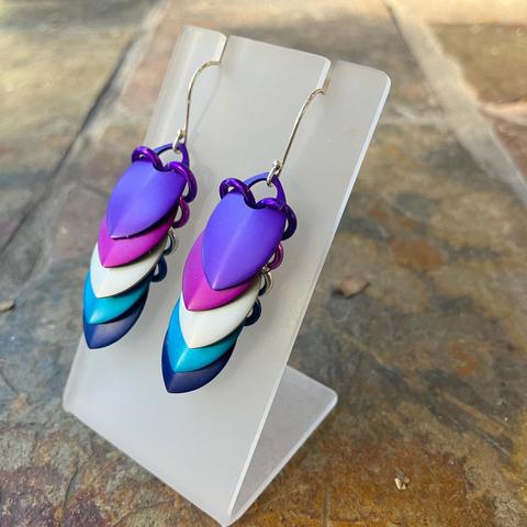 A pair of scalemail earrings on a white display stand. The earrings each consist of five overlapping 'scales' - top one dark purple, next violet, white-silver, light blue, then finally dark blue. They're very pretty.