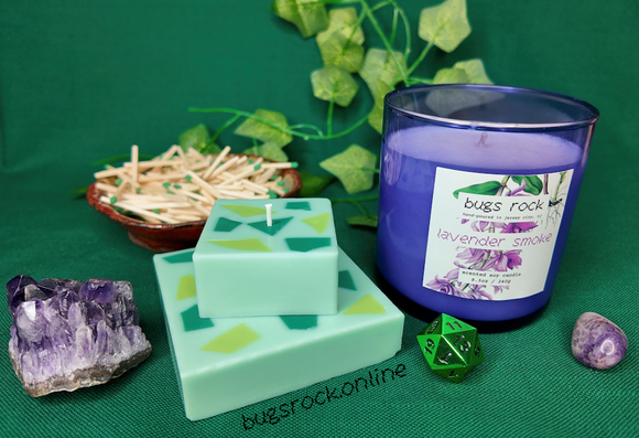 A photo of two candles on a green backdrops surrounded by amethysts, strawberry candies, a dish filled with green-tipped matches, and a green chrome 20 sided roll to 11 because my birthday is on the 11th. The candle on the left is a green two-tiered cubic pillar candle with a handmade terrazzo-like style green pattern on the tops of each tier. on the right is a periwinkle glass with white wax candle called lavender smoke.