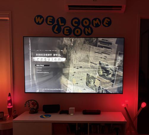 A photo of my wall-mounted TV. The splash screen for the game Resident Evil 9 is displayed. Surrounding the TV are multiple red tinted lights and a “Welcome Leon” sign