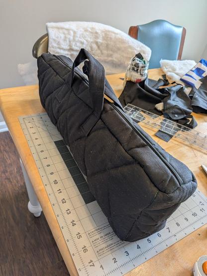 A closed bag. It is a homemade black instrument case for an alto flute. It's about 20 inches long, 7 inches high, and 4 inches wide. It is made of plain black fabric and quilted on the sides. It sits on a messy kitchen table with lots of sewing items scattered around.
