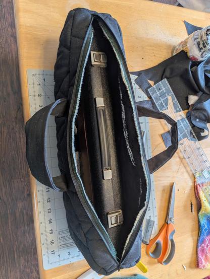 An open bag. It is a homemade black instrument case for an alto flute. It's about 20 inches long, 7 inches high, and 4 inches wide. It is made of plain black fabric and quilted on the sides. It sits on a messy kitchen table with lots of sewing items scattered around.