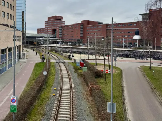 A spur track going underneath the viewer, with Amersfoort station in the background, and bikes parked to the side. The scene has a lot of paved roads curving around, and greenery in between. In the background, a large modern brick building