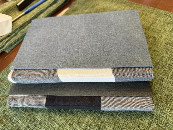 Two stacked fabric-covered notebooks are displayed on a green textured surface. The top notebook has a solid blue-gray cover, while the bottom notebook features a multi-colored pattern on the edges, including stripes and solids in various shades.