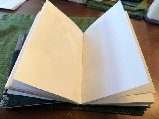 An open blank notebook rests on a textured green surface. The visible pages are clean and empty, with a slight angle revealing the binding and edges. The background features additional items, including a dark-colored cover partially showing beneath the notebook.