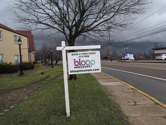 A posted real estate sign has been replaced with a sign board that says, "Book a tour today - It's free!" Also the Bloop Museum logo and website, bloopmuseum.com
