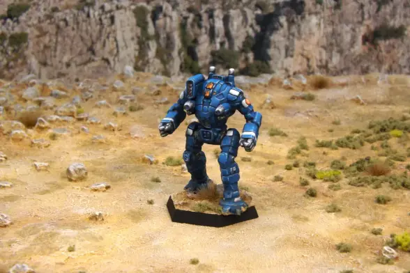 A painted miniature of an Ostroc 'Mech.  It is blue with white highlights.  The cockpit glass is orange.  The emblem of the 1st Aragon Borderers, a blue crenelated shield, is on the right knee.  The sunburst emblem of the Federated Suns is on the left arm.  The number "70" is on the lower left leg.  The miniature is photographed on rocky terrain.