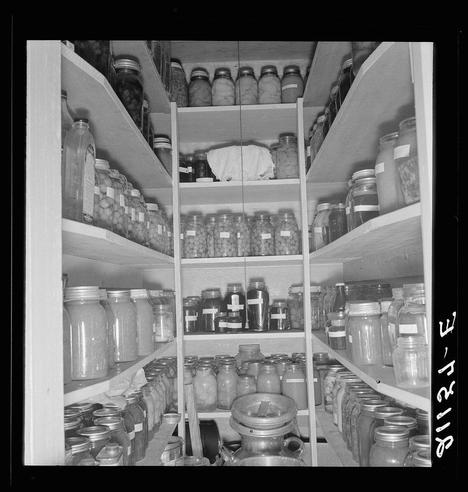 The image depicts a well-organized pantry filled with numerous jars of preserved food. The shelves are neatly arranged, showcasing an assortment of items such as pickles and jams in clear glass jars. Labels on some of the jars indicate different contents or possibly production dates. This storage space is indicative of careful preservation practices, likely aimed at extending the shelf life of perishable goods for consumption during times when fresh produce might be unavailable.

The black-and-white nature of the photograph suggests it could be from a past era, reflecting traditional methods of food conservation before modern refrigeration techniques became widespread. The organized arrangement and variety of preserved foods highlight an approach to self-sufficiency in sustenance that is both practical and resourceful.