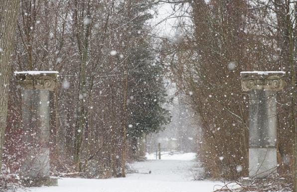 Two stone columns mark the entrance to a trail through woods.  The ground is white and big snowflakes are falling.