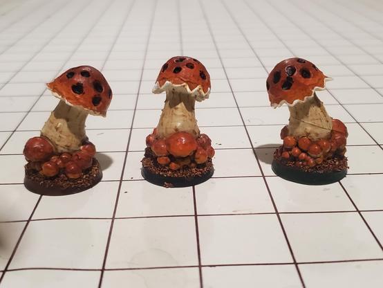 3 mushroom enemies for DnD on a square grid playmate. They model itself is extremely phallic looking, but the painted minis look appropriately like mushrooms. They have orange brown caps with spots and dirty off-white stems. They're surrounded by smaller mushrooms if similar coloring. The stems are particularly well done, and have the striated, faintly brown vertical textures that real mushrooms often have.