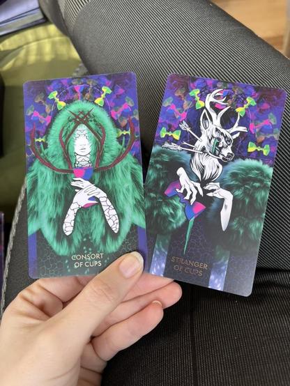 consort of cups next to the stranger of cups. both cards are extremely vibrant colors and a combo of digital collage and textures and illustrations. The consort wears vibrant furs and holds antlers in front of aer face with cracked hands. The Stranger has the head of a deer and with claw-like hands pours out a chalice