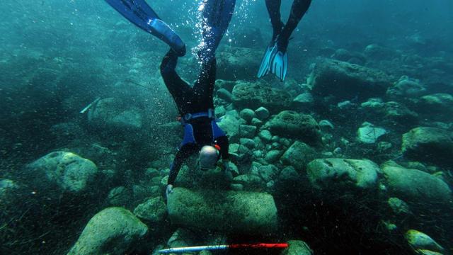 A diver in scuba gear and blue fins is upside down in clear water, examining a large rectangular stone on a rocky seabed while another diver with blue fins hovers nearby; an archaeological measuring stick lies on the bottom in front of the stone.