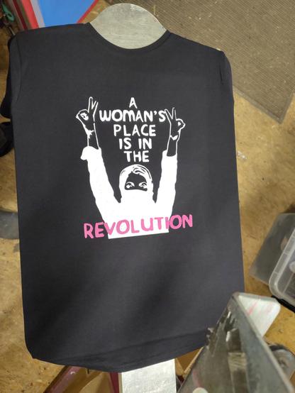 schwarzes Shirt auf der Siebdruckmaschine: zweifarbiger Druck, in weiß eine vermummte Person, die die Hände in die Luft reckt und der Text "a woman's place is in the" darunter in "pink" (der Name der Farbe ist Fuchsia) das Wort "revolution"
