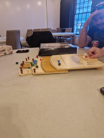 View of someone's hand flicking a die up a small wooden board on a table. The board has various multiplier sections, with the highest scoring ones as tabs at the top, and there are already a few different coloured dice on it from the other players.