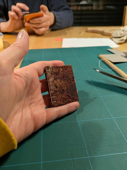 Hand holding a mini book showing the front. It's bound in a brown toned jacquard fabric with woven patterns