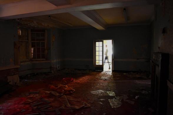 A haunting view of a decaying room where a single burst of light from a doorway cuts through the shadows, framing a lone figure walking across the bright exterior.