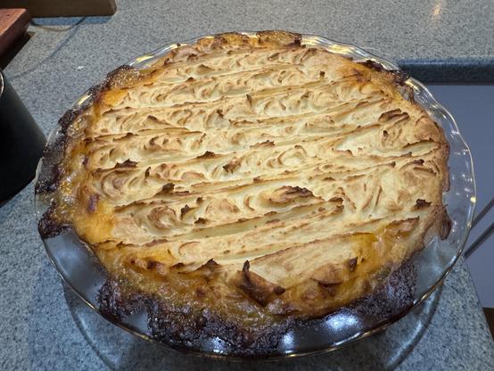 A pyrex pie dish full of patterned mash potato that has browned and crisped on the peaks. Underneath but not visible is a layer of beef mince with carrots, onions, garlic, mushrooms and butter beans.