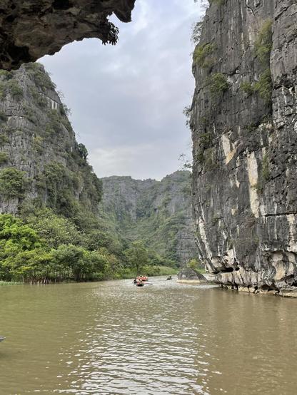 A photo of a river flowing between high, almost vertical cliffs. The water is calm and muddy. A few rowing boats with tourists are floating along. There is some forest along the coast and in the cliffs. The sky is partially cloudy.
