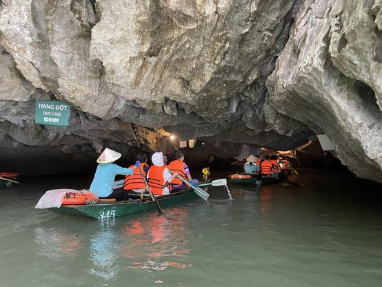 A photo of several rowing boats with tourists floating into a cave. Each boat is steered by a local with a Vietnamese hat and a uniform. On the cliff near the entrance the sign reads: Hang Dot - Dot cave - 1000 m. The cliffs of the cave are low lying and very rough. The water is murky.