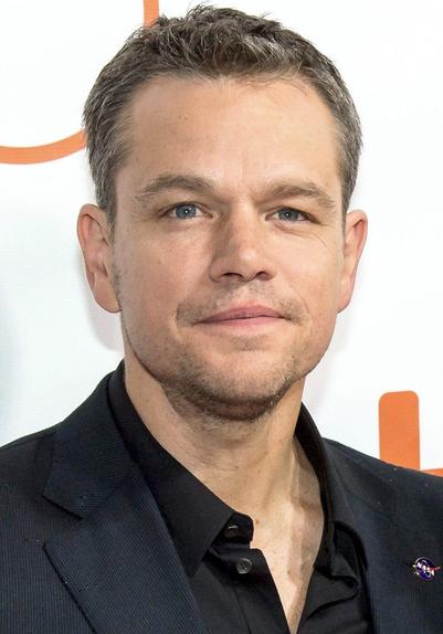 Matt Damon steht vor einem grauen und orangefarbenen Hintergrund. Er trägt ein schwarzes Jackett und ein schwarzes Hemd. Der Mann lächelt leicht und hat einen stoppeligen Bart. An seinem Jackett befindet sich ein Pin mit dem NASA-Logo.