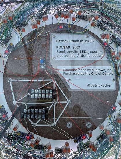 Reverse side of the artwork. An upright metal ring with lots of wires and electronic components attached, running outward. Inside there's a small plaque:
Patrick Ethen (b. 1988)
PULSAR, 2021
Steel, acrylic, LEDs, custom electronics, Arduino, code
Commissioned by Midtown, Inc. 
Purchased by the City of Detroit