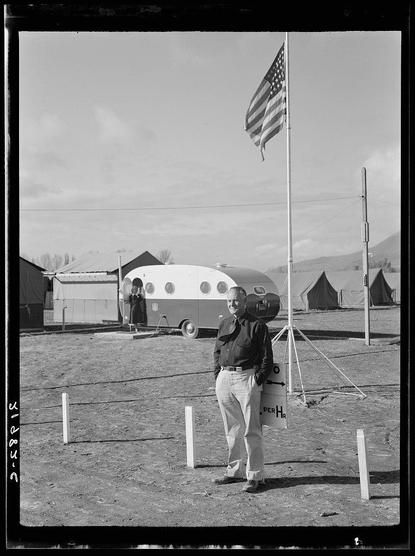 This black and white photograph depicts a scene at what appears to be an outdoor campsite or temporary settlement. In the foreground, there is a man standing beside a flagpole bearing an American flag. He wears a dark jacket, light-colored trousers, and has his hands clasped in front of him while holding a sign that reads "PER HR" which suggests he may be managing time-based services at this campsite.

Behind him are two portable trailers; one is white with rounded edges and the other appears to have windows. The setting includes several small buildings or tents pitched on flat ground, indicating some form of organized settlement or temporary housing arrangement.

The environment looks like an open field under a partly cloudy sky with visible mountains in the distant background, contributing to a sense of vastness around this campsite. There is no indication of any specific activities taking place at that moment captured by the photo; it serves more as a snapshot of life within such settlements during the period.

The photograph seems to be part of a collection documenting rural or migrant laborers' lives and work, possibly from an era where agriculture required temporary housing for seasonal workers. The presence of portable trailers suggests mobility and transience in living arrangements typical for agricultural communities.

For further context on this image's origin,  [...]