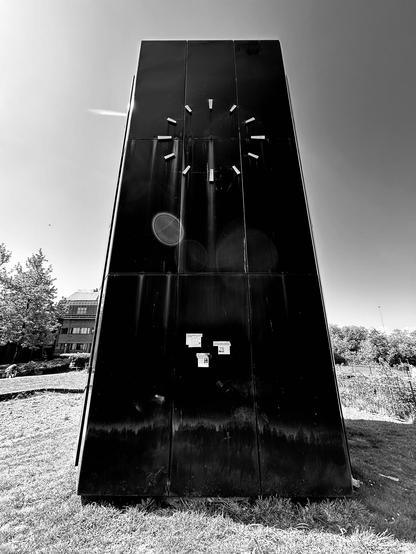 An ominous clocktower in black cladding and featuring a numberless, handless clock face. The structure is stained and has some sort of tidemark near its base. Someone has attached three newspaper clippings to the front, all with a bizarre conspiratorial theme. A blob-like lens flare artefact appears near the centre of the image. The Civic Centre Clock, San Francisco Libre Walk, Reading, Berkshire, UK. Photo by Gearóid Burke.