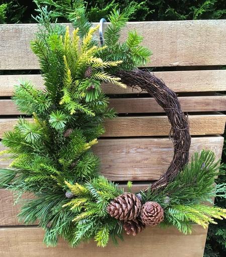 A natural wreath made of fresh green evergreen branches and pine cones on a brown grapevine/morning glory vine base, hanging against a wooden fence.