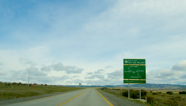 under a massive sky full of sketchy white and grey cirrus and other clouds, a road stretches into the distance, where low mountains rise along the horizon. a road sign tells the viewer this is the "Ruta del Fin del Mundo" and includes a very basic map above a final sign reading Cirucito Baquenos