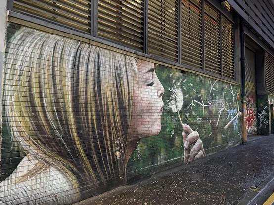 A large mural depicting a woman with long hair blowing on a dandelion, surrounded by whimsical green foliage. The artwork blends realism with a dreamy atmosphere, capturing the moment as dandelion seeds disperse into the air.