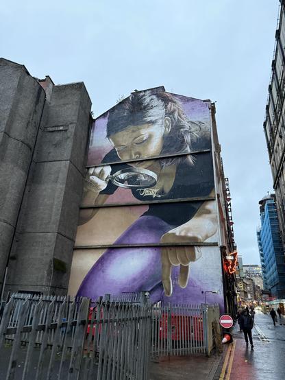 A large mural on the side of a building depicts a woman examining something through a magnifying glass. She has long hair and is wearing a black top with purple pants. The image features urban surroundings, including a street with barriers and a red stop sign