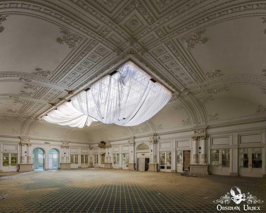 An ornate, abandoned hotel ballroom features white and gold decor with a draped skylight diffusing light. The vast room has a patterned floor.