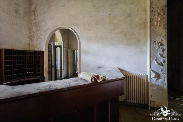 An abandoned hotel reception desk features a dark wooden counter, a key rack, and an archway. Peeling paint covers the decaying wall