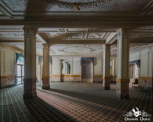 An opulent, abandoned hotel ballroom with ornate ceilings, grand pillars, and patterned carpet. Sunlight casts shadows.