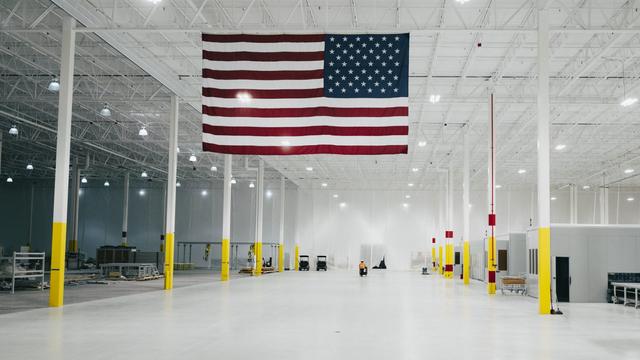 An expansive, empty warehouse interior featuring a large American flag hanging from the ceiling, yellow painted columns, and various industrial equipment scattered throughout the space. The bright lighting illuminates the clean, white floor. An expansive, empty warehouse interior featuring a large American flag hanging from the ceiling, yellow painted columns, and various industrial equipment scattered throughout the space. The bright lighting illuminates the clean, white floor.
